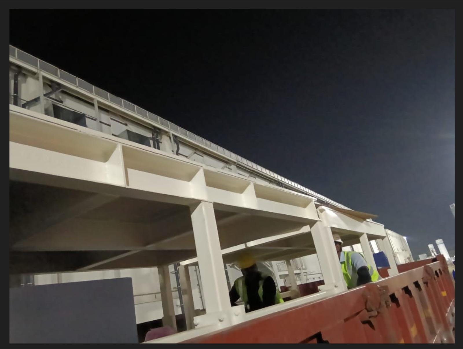 Elevated fabricated steel walkway painted white, viewed from below during a night installation with workers in hi-vis vests