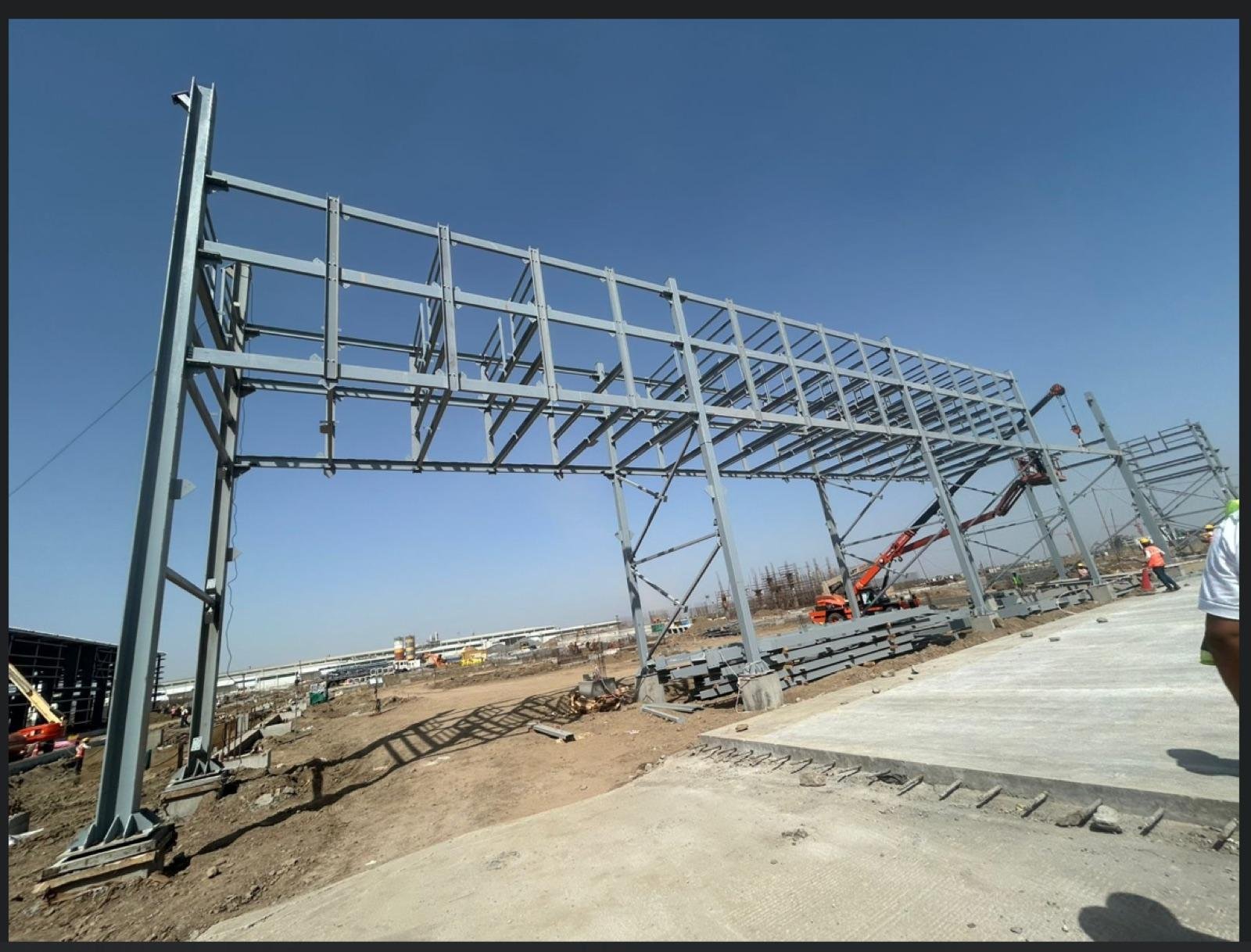 Structural steel portal frame under erection at an industrial site with a mobile crane and clear sky
