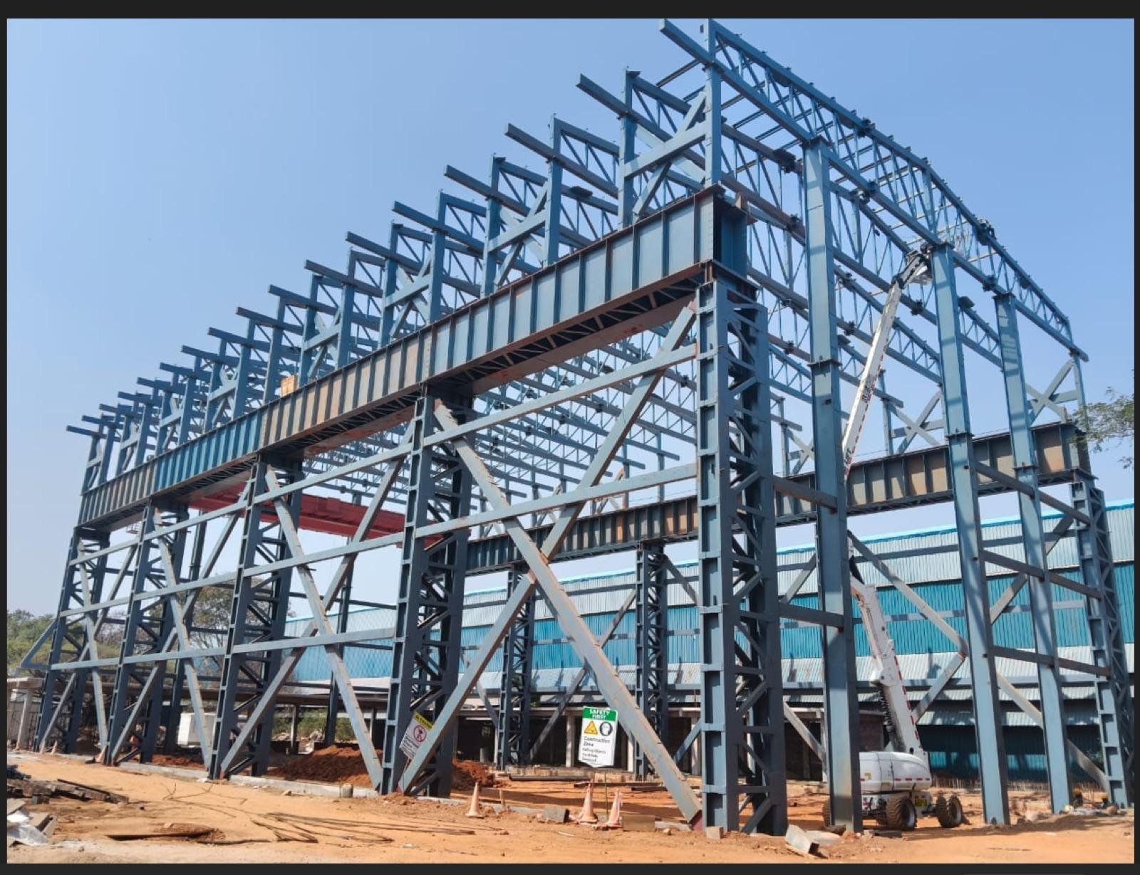 Tall blue-grey steel portal frame structure under construction at an industrial plant, with a boom lift alongside