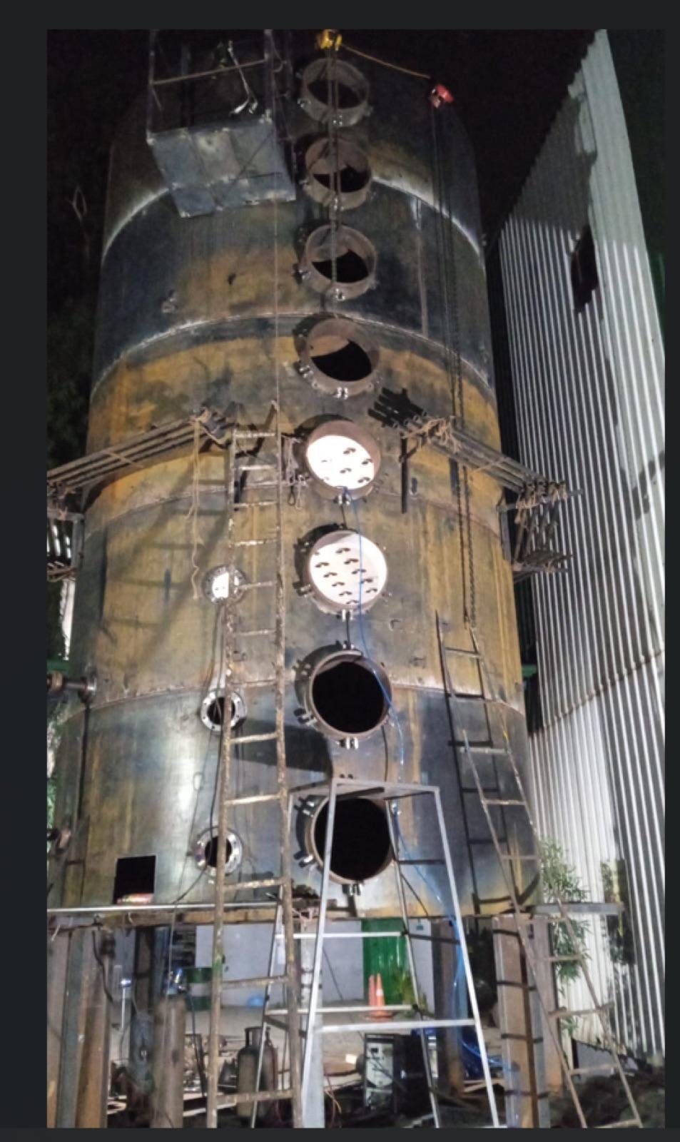 Tall fabricated steel process vessel with a row of circular nozzle openings, surrounded by access ladders and scaffolding, lit at night