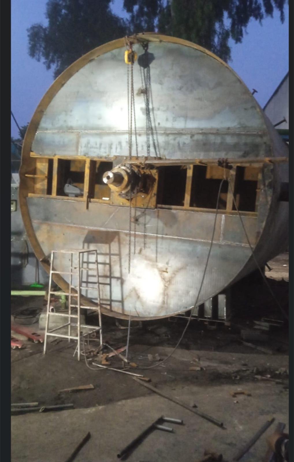 Large cylindrical steel tank under fabrication in a yard at dusk, rigged to a chain hoist with an access ladder